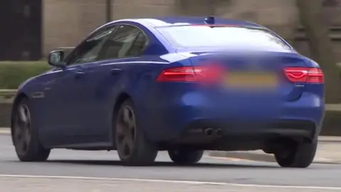 A blue saloon car driving down a road. Its number plate has been blurred.