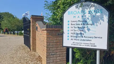 Debbie Tubby/BBC A white sign in front of an entrance to a marina. The sign is white and says, Welcome to BROADS EDGE MARINA. There is a gate at the front of the entrance which is open. Boats in the water are behind some trees in the background.