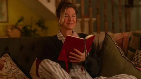 Universal Studios Renee Zellweger as Bridget Jones in the fourth Bridget Jones film. She is dressed in white and blue striped pyjamas with a grey cardigan. She is sat on a sofa holding a red diary and smiling. 