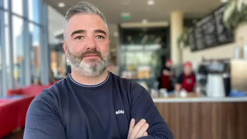 Cormac McAteer wears a grey sweatshirt. He has a grey beard and grey hair. Relish cafe is behind him, with staff working behind the counter. 