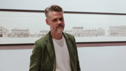 A stylish middle-aged man with a short greying beard and gelled hair poses in front of a large drawing of a streetscape.