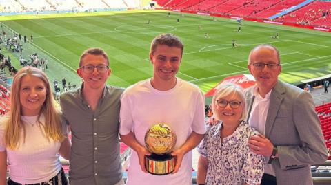 JJ Lacy holding golden ball trophy with four members of his family, in the stands at Wembley with the pitch behind them