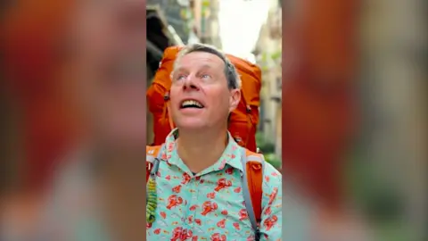 A man wearing a turquoise shirt with red lobsters on and an orange backpack looks up to the sky as he walks down a street.