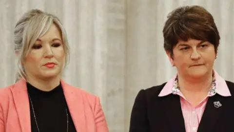 Michelle O'Neill standing next to Arlene Foster. O'Neill has blonde hair tied back with a fringe wearing a pink blazer and black top, and a long silver necklace. She is wearing makeup with pink lipstick. Foster has short brown hair and is wearing a pink striped shirt with a black blazer.