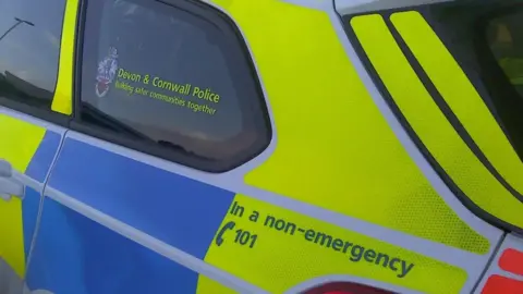 A luminous yellow and blue Devon and Cornwall Police car. It says: In an emergency call 101.