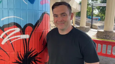BBC A man with short brown hair and a black T-shirt is smiling while standing next to a pillar painted blue with a red poppy design.