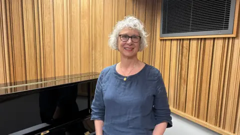 Katy Lambert/BBC A woman with white curly hair, black-rimmed spectacles and a blue top smiles into the camera. She is stood next to a piano and a wood-panelled wall is behind her.