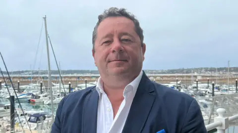 A man with a white shirt under a navy blue suit standing in front of the quay.
