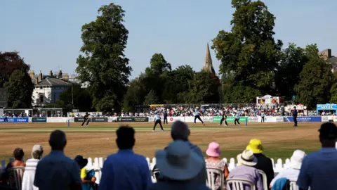 Getty Images/Dan Istitene A cricket pitch with spectators sitting on plastic chairs in the foreground and a bank of spectators on the opposite side. There are several tall trees in the background and a church spire against a blue sky