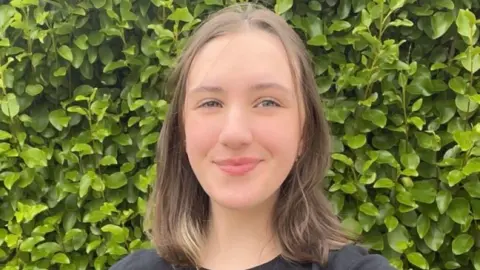 Ciara Murray Girl with shoulder length brown hair and black top standing in front of green hedge