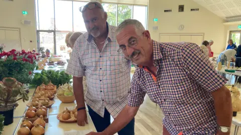 Stuart Smale on the left is wearing a white, blue and orange chequered shirt with glasses on his head. Geoff Gallienne on the right is wearing glasses and a blue and red chequered shirt. Both are standing next to a display of onions.