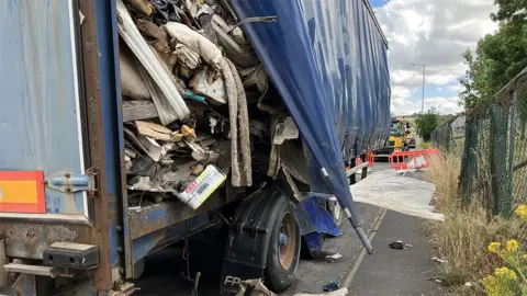 BBC View of the side of the large HGV trailer, covered in blue plastic, with a triangular-shaped opening to the side where rubbish is piled high and falling out on to the pavement