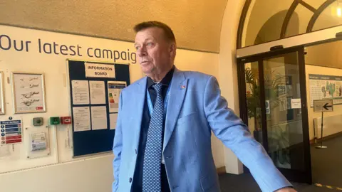 Local Democracy Reporting Service A man in a blue suit and tie looking to the side in a hallway with a notice board on the wall behind him.