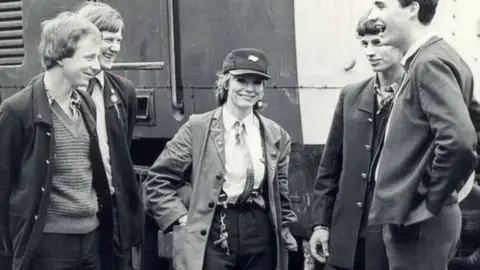 Aslef A black and white photo shows a young woman in a train driver's uniform standing in front of a train flanked by men