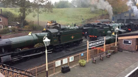 A train from the Severn Valley Railway in Bridgnorth station. The train is dark green and beyond it part of a house can be seen and a green field. A train platform is visible which has a lamppost on, light pint railings and a sign saying 'Bridgnorth'. 