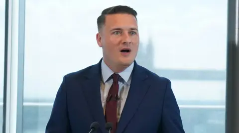 Wes Streeting wearing a navy suit and a red tie. He is standing at a lectern on front of a view of Tower Bridge in central London