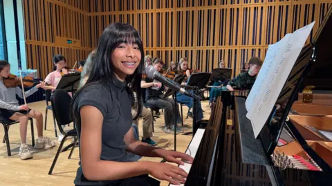 Skye playing a grand piano with the Young Northern Sinfonia Orchestra inside The Glasshouse. She is wearing a black dress and has long black hair. She is smiling at the camera while playing.