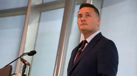 Getty Images Wes Streeting stood at a lectern delivering a speech. He is wearing a navy blue suit, pale blue shirt, and red tie. 