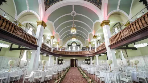 The Grand Palace Inside the building. There are white chairs and tables and a carpet which has flowers down the sides. There is a brown balcony and arched ceiling and pillars which are white with gold trim. The ceiling is arched and there are two chandeliers in the centre. 