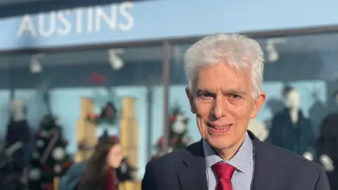 David Austin, wearing a blue and white fine striped shirt, a red tie and a blue suit jacket and standing outside Austins with the store behind in a blurred background.
