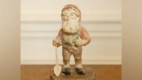 Lamport Hall A garden gnome whose paint has faded. He has a beard and has a spade in his right hand. 