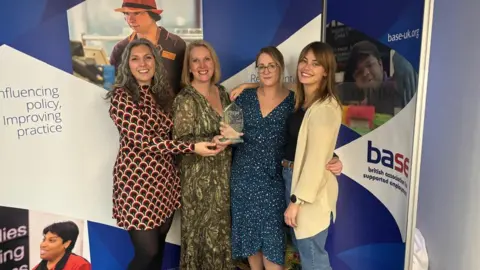 Elaine Strappini Left to right is Alexandria Martinson wearing a red and black patterened dress, Elaine Strappini is wearing a green and white dress, Claire Smith is wearing a blue and white dress and Rebecca Mauger jeans, a black top and a white cardigan. They are stood in front of a blue and white sign. 