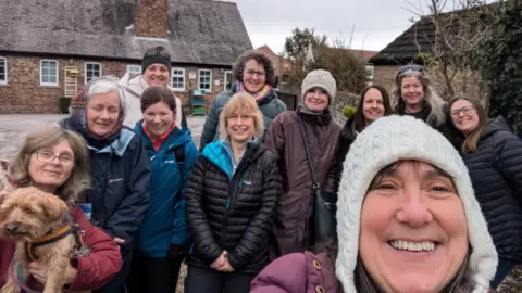 Provided A group photo of 11 women in coats and a dog on a walk. Some are also wearing woolly hats.