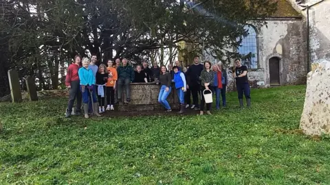 Ruth Marshall A large group of people gather in the churchyard around a concrete tomb, there are graves in the background as well as trees and a section of the church can also be seen