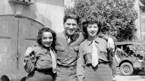 Hestercombe House Trust A black and white image of a man in a shirt, with his arms around two women, who are both in a uniform comprising shirts, ties and skirts. They stand in front of stable doors, with a buggy-style vehicle in the background.