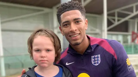 Bev Price A boy with long hair stands with footballer Jude Bellingham dressed in his England tracksuit. They are stood outside a football training facility.