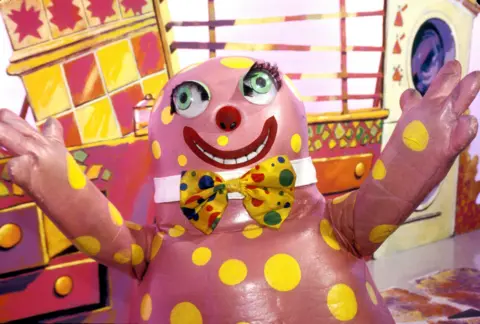 Mr Blobby wears a yellow polka dot bow tie and has his arms raised to the camera.