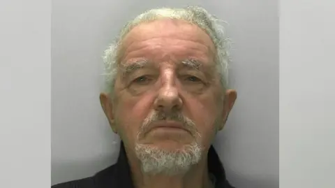 Gloucestershire Police A custody image of Philip Wood. He has coarse, curly white hair cut short, a white goatee and thick grey eyebrows. 
