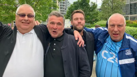 Lewis Adams/BBC Ray Bentley is wearing a blue Southend top and navy Southend jacket. He has his arm around a man with brown hair. Ray is on the left wearing a white T-shirt and black jacket. He has an arm on the shoulders of a man wearing all black and cheering.