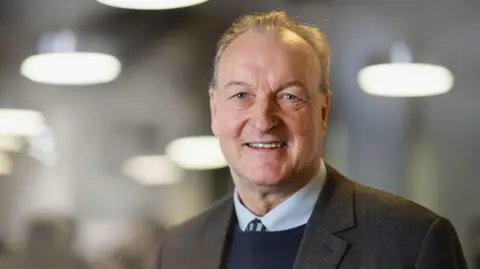 A man with grey hair in a grey suit. Under his jacket is a navy jumper over a pale blue shirt and tie