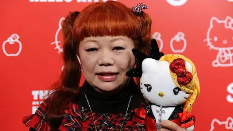 Getty Images A medium close-up of Yuko Yamaguchi wearing a red and black tartan dress holding a Hello Kitty doll who is wearing a sparkly red and black outfit
