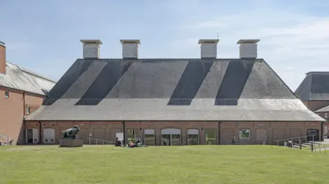 Timothy Soar A building at Snape Maltings Concert Hall, in the foreground is a patch of grass with a red brick building behind it, it has a large sloped dark roof with four vents on top.