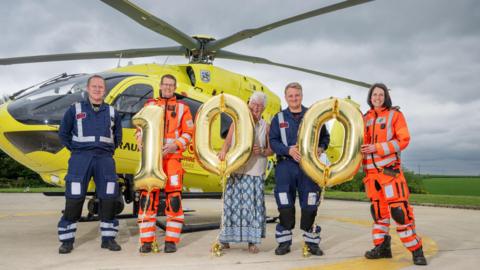 Janet Swallow stands with Yorkshire Air Ambulance crew and 100 balloons in front of a helicopter