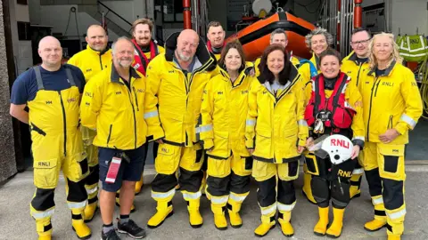 RNLI / Chris Page and Lisa Newberry Jones and Speirs standing in the middle alongside eleven volunteers from the RNLI. They are all wearing yellow uniform and are stood in front of the lifeboat station. 