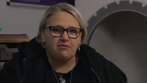Childminder Victoria Cook. She has short, blonde hair, glasses and is wearing a black zip-up jacket and has silver necklaces on. In the background is a mirror.