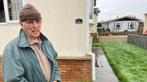 James Grant/BBC David McGovern, wearing a brown polo shirt, a blue coat stands and a brown wollen hat, stands next to his property. A path on the right hand side is covered in water.