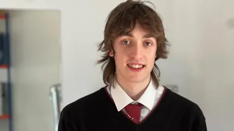 Maxime in his school uniform, a black sweater, white shirt and a red tie.