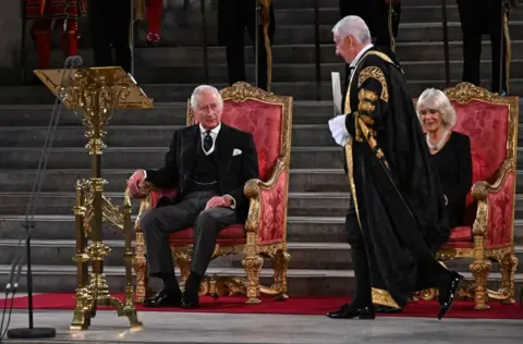 BEN STANSALL/AFP Speaker of The Commons Lindsay Hoyle walks past Camilla, Queen Consort to hand a document to King Charles III