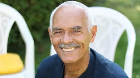 Charles Todd smiles wearing a navy blue shirt and against the backdrop of two white chairs