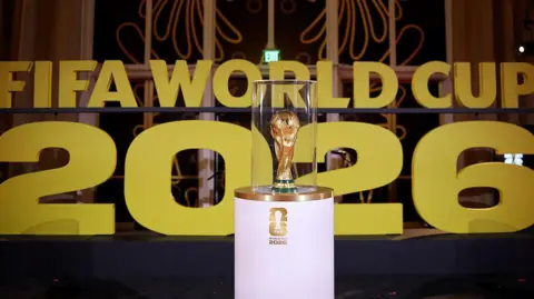 The World Cup trophy on display on a white stand prior to the draw in December in Washington with Fifa World Cup 2026 in big gold letters in the background.