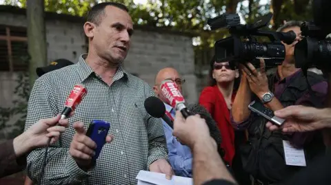 Getty Images Cuban activist Jose Daniel Ferrer speaking to journalists in 2015. Several journalists are standing around him with mics as he speaks. One is holding a camera. 