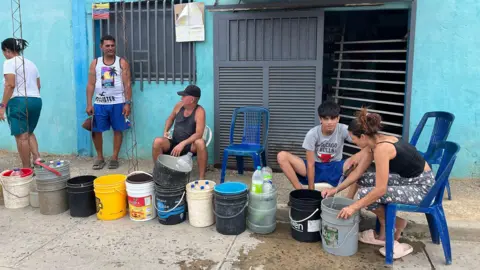 Two men, two women and a boy queue for water in a street in Cumaná. They have lined empty buckets up. Some are sitting on plastic chairs while others are are standing. Some buckets contain empty plastic water bottles waiting to be filled.
