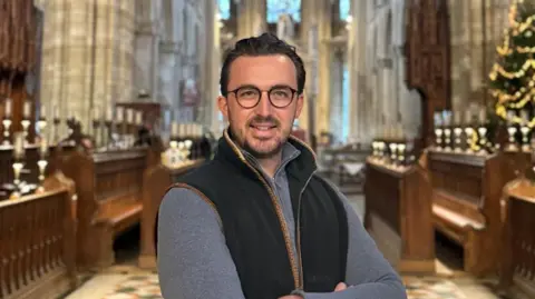 Peterborough Cathedral Jack Pishhorn, a man, wearing glasses, a grey full-sleeve top under a navy sleeveless fleece, and he is smiling for the camera. He is standing in front of what looks like the choir's pews in a cathedral. There is a decorated Christmas tree on the right.