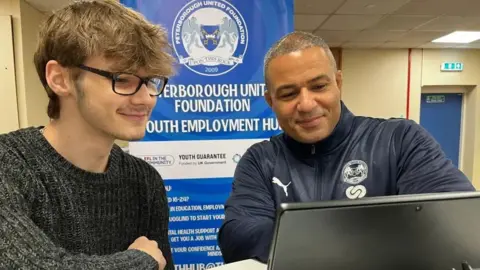 Young man wearing a grey jumper and glasses looking at a laptop with an older man with information about the employment hub behind them.