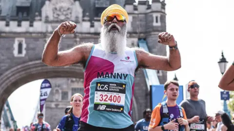 Manny Singh Kang A man running over Tower Bridge as part of the London Marathon. He has a white, pink and blue vest on, with the Dementia UK charity name on it. He has a race number attached to it with the number 32400 on it. He is wearing a yellow head scarf and sunglasses and has both arms up, flexing