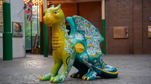 A large dragon sculpture that is green, yellow and blue, with yellow claws. It has drawn stickers of people on it, with a yellow circular logo that reads "Compton Care"